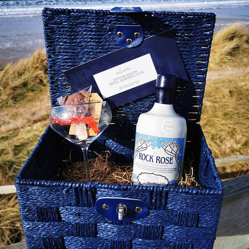 Content of the Refill Rewards Club Gift Set Hamper including a bottle of Rock Rose Gin, a branded glass, a Caithness Chocolate Mr Mackintosh Lollipup, a gift voucher for our Refill Rewards Club in a blue hamper